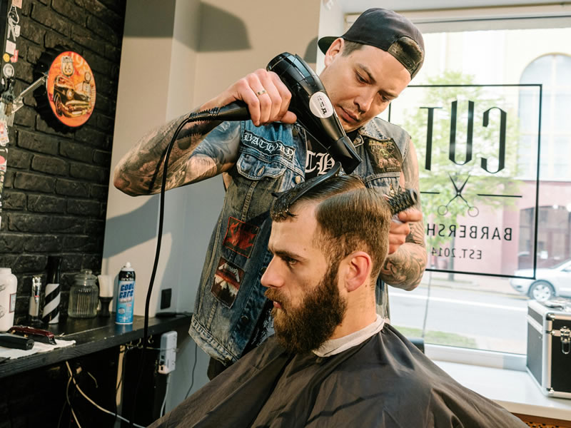 Taglio uomo 2026: barbiere asciuga i capelli con phon. Tendenze capelli uomo e cura della barba nel barber shop.
