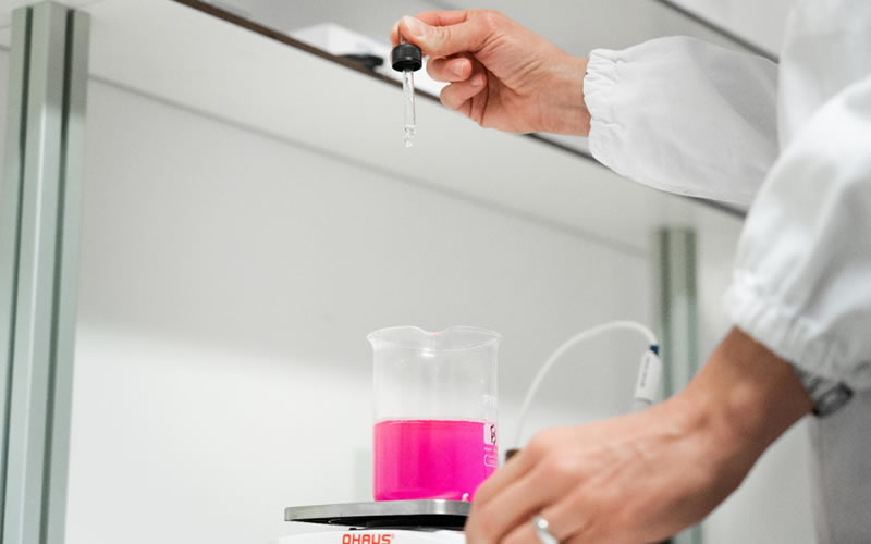 Creazione prodotti professionali capelli Dott. Solari: scienziato aggiunge liquido con pipetta a beuta rosa. Laboratorio cosmetico avanzato.