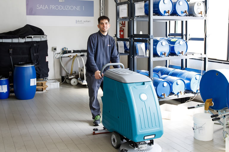 Dott. Solari: addetto alla pulizia industriale con lavasciuga pavimenti professionale in area di produzione. Barili blu sullo sfondo.