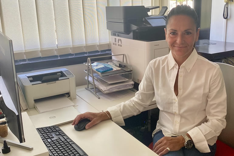 Chiara siede alla sua scrivania in ufficio, sorridendo verso l'obiettivo mentre utilizza il mouse del computer tra stampanti e documenti.