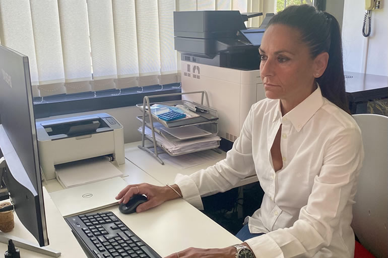 Donna con camicia bianca lavora al computer in ufficio, mano sul mouse, stampante e documenti sulla scrivania.