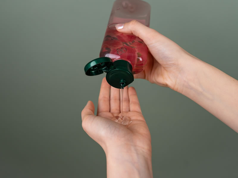Mano che versa del gel detergente trasparente da un flacone rosato con tappo verde su uno sfondo color salvia.
